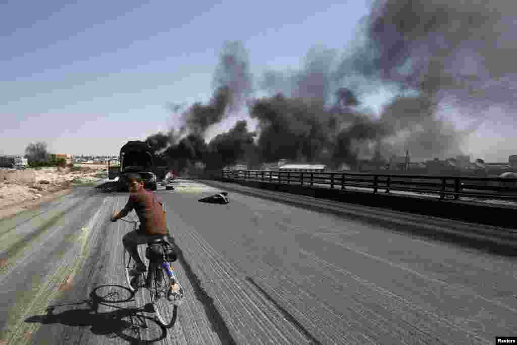 A Syrian boy rides a bicycle as smoke rises over the Syrian city of Aleppo after missiles fired from a fighter jet hit petrol tankers in the Bab al-Nayrab district August 27, 2012. 