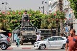 FILE - Tunisians walk past a military armored personnel carrier at Avenue Habib Bourguiba in Tunis, July 30, 2021. Political turmoil in Tunisia has left its allies in the Middle East and West watching to see if the fragile democracy will survive.