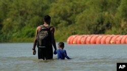 ARCHIVO: Dos migrantes caminan cerca de unas grandes boyas usadas como barrera fronteriza flotante en el Río Grande, o Bravo, el 1 de agosto de 2023, en Eagle Pass, Texas.