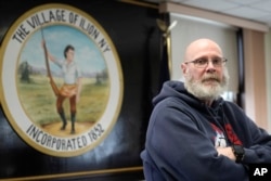 Ilion Mayor John Stephens poses for a picture in front of the seal of the village at the municipal building in Ilion, N.Y., Thursday, February 1, 2024. The seal features Eliphalet Remington, the founder of Remington Arms Co. (AP Photo/Seth Wenig)