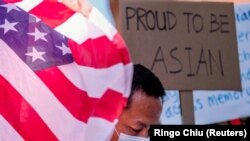 Seorang demonstran berdiri di antara bendera AS dan sebuah poster saat unjuk rasa menentang kejahatan kebencian terhdapa warga Asia-Amerika di luar balai kota Los Angeles, California, Sabtu, 27 Maret 2021. (Foto: Ringo Chiu/Reuters)