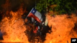 Seen through a burning street barricade, an anti-government demonstrator waves a Chilean flag in Santiago, Chile, Oct. 28, 2019.