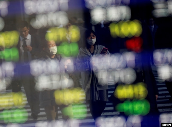 Pedestrians wearing facial masks are reflected on an electric board showing stock prices outside a brokerage at a business district in Tokyo, Jan. 30, 2020.