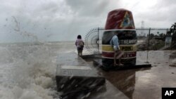 FILE - Tourists pose next to the Southernmost Point marker as large waves crash on the sea wall, July 8, 2005, in Key West, Fla., as winds from Hurricane Dennis stir up sand and surf. 