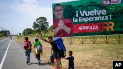 Una familia de venezolanos que emigra a Colombia hace autostop frente a una valla publicitaria que invita a votar por el candidato a gobernador del partido gobernante, Jorge Arreaza, en Barinas, Venezuela, el viernes 21 de enero de 2019. 