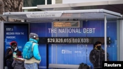 People wearing protective face masks wait at a bus stop with a display of the current national debt amid the COVID-19 pandemic in Washington, Jan. 31, 2022. 