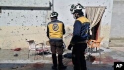 In this photo provided by the Syrian Civil Defense White Helmets (SCDWH), which has been authenticated based on its contents and other AP reporting, SCDWH workers stand at the site where a shell struck in the Maaret al-Naasan village in Idlib province, Syria, Feb. 12, 2022.