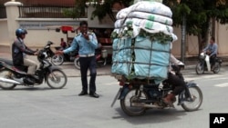 A Cambodian man, right, drive his motorbike overloaded with vegetable sacks as he passes a traffic police officer, center, in Phnom Penh, Cambodia, Tuesday, Aug. 25, 2015. (AP Photo/Heng Sinith)