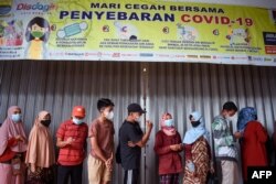 Warga berdiri di bawah spanduk yang mengingatkan warga untuk memakai masker dan tips lainnya dalam mencegah penyebaran COVID-19 saat antre membeli minyak goreng di Bandung,18 Februari 2022. (TIMUR MATAHARI / AFP)
