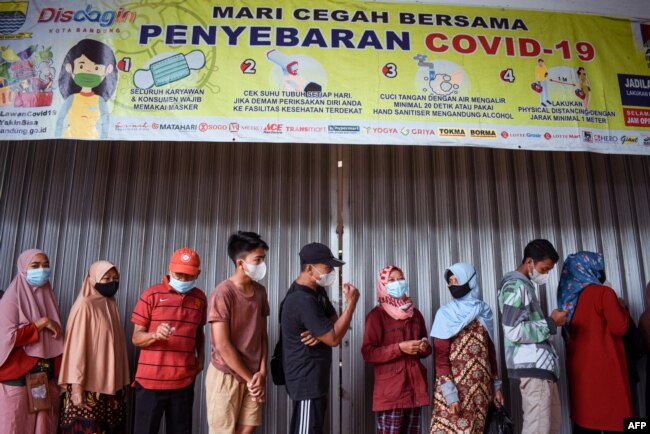 Warga berdiri di bawah spanduk yang mengingatkan warga untuk memakai masker dan tips lainnya dalam memerangi Covid-19 saat mereka mengantri untuk membeli minyak goreng di Bandung pada 18 Februari 2022. (Foto: AFP)