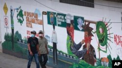 Dua pria mengenakan masker berjalan melewati mural bertema virus corona di Medan, Sumatera Utara, Kamis, 17 Februari 2022. (AP/Binsar Bakkara)