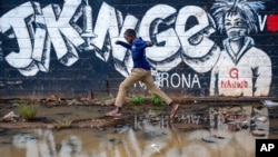 FILE - A boy leaps across a flooded area next to an informational mural with words in Swahili advising people to protect themselves from the coronavirus and get vaccinated, in the low-income Kibera neighborhood of Nairobi, Kenya, June 12, 2021. 