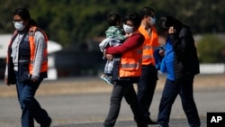 FILE - In this March 12, 2020 file photo, an immigration worker in an orange jacket and wearing a mask as a precaution against the spread of the new coronavirus, carries a young Guatemalan migrant who was deported from the U.S.