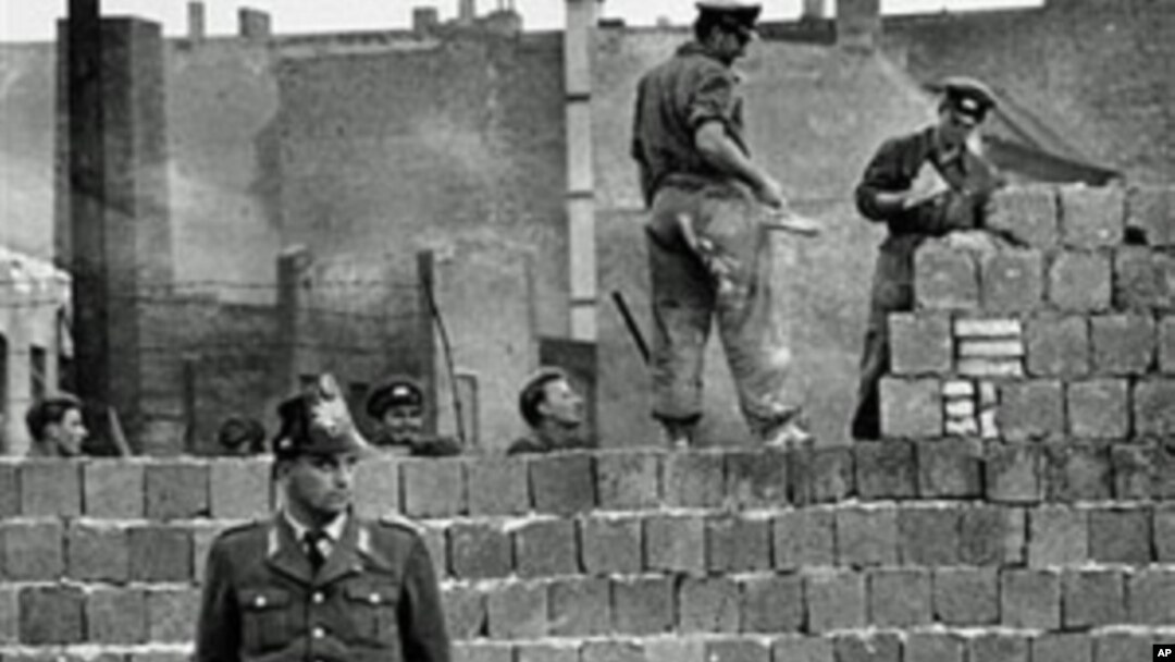图为1961年10月7日一名西柏林士兵站立在分割东、西柏林的混凝土墙边。他身后为东柏林工人正在加高围墙