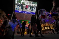 Shoppers walk by a woman selling balloons near a giant TV screen broadcasting a news of demonstrators protest against the U.S. election results, at a shopping mall in Beijing, Nov. 8, 2020.