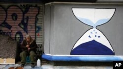 A shoe shiner tries to keep warm next to an hourglass graffiti in Athens, Greece, Feb. 20, 2012.