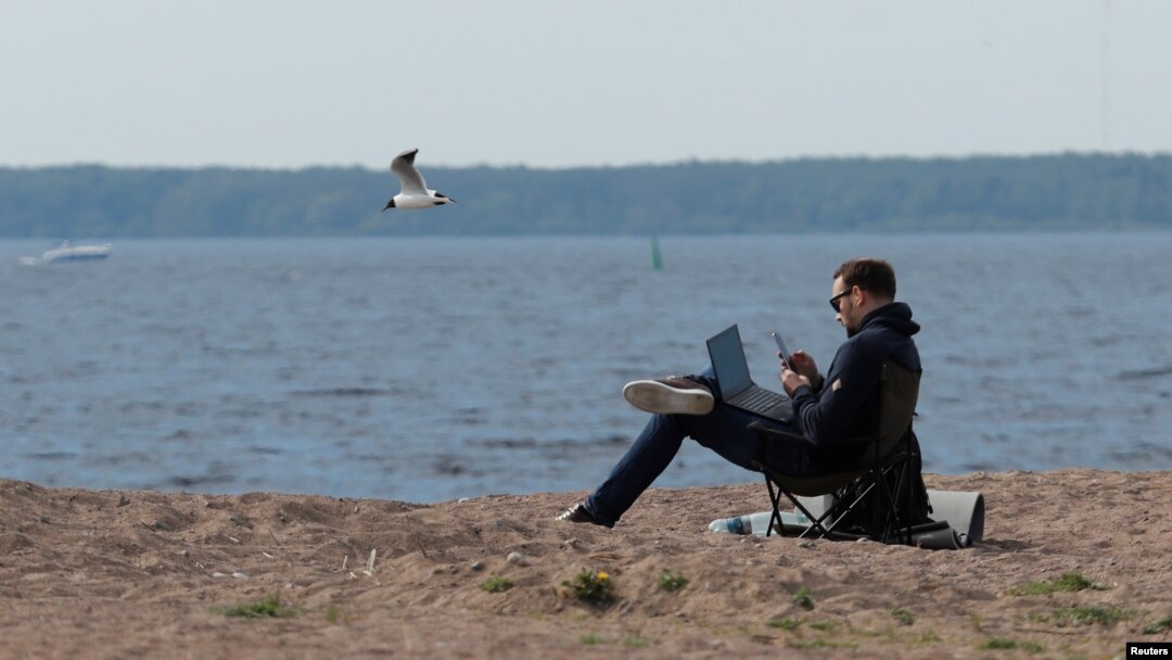 Seorang pria bekerja dengan menggunakan smartphone dan laptopnya, sambil duduk di pantai Teluk Finlandia di tengah pandemi Covid-19 di Saint Petersburg, Rusia, 27 Mei 2020. (REUTERS / Anton Vaganov)
