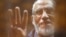 FILE - The Brotherhood's Supreme Guide Mohamed Badie waves with the Rabaa sign, symbolizing the support of the Muslim Brotherhood, as he stands behind bars during his trial with ousted Egyptian president Mohamed Morsi and other leaders of the Muslim Broth