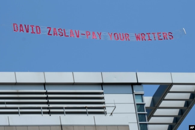 A message in support of the Hollywood writers' strike is pulled by an airplane in the sky above Boston University commencement ceremonies, Sunday, May 21, 2023, in Boston, during an address by David Zaslav, president and CEO of Warner Bros. Discovery. (AP Photo/Steven Senne)