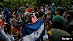 Migrantes de Honduras sostienen una bandera hondureña mientras están parados frente a soldados guatemaltecos que bloquean una carretera para evitar que los migrantes lleguen a la frontera de México, en San Pedro Cadenas, Izabal, Guatemala, octubre 2020.
