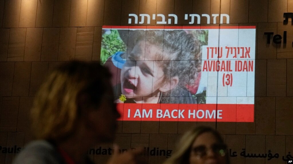 2023年11月26日，人们走过特拉维夫一栋大楼上投射出的4岁阿比盖尔·伊丹(Abigail Edan)的照片，她是哈马斯扣押的人质，于周日前夕被释放。伊丹的父母均被哈马斯武装分子杀害。10月7日，她遭到绑架，这是一次跨境袭击，促使以色列向哈马斯宣战。 （美联社照片）(photo:VOA)