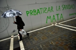 A woman takes shelters from the rain under an umbrella while passing graffiti reading,''Privatizing Health Service kills. Capitalvirus'', in Pamplona, northern Spain, March 16, 2020.