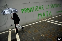 A woman takes shelters from the rain under an umbrella while passing graffiti reading,''Privatizing Health Service kills. Capitalvirus'', in Pamplona, northern Spain, March 16, 2020.