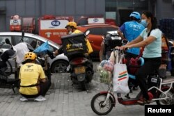 A woman wearing a face mask rides an electric bicycle with her groceries past delivery workers of Meituan and Ele.me, in Beijing, China, July 13, 2020.