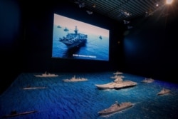 A TV screen showing the U.S. Navy fleet sail in formation near the models of Liaoning aircraft carrier with navy frigates and submarines on display at the military museum in Beijing, Aug. 1, 2019.