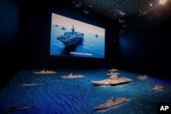 A TV screen showing the U.S. Navy fleet sail in formation near the models of Liaoning aircraft carrier with navy frigates and submarines on display at the military museum in Beijing, Aug. 1, 2019.