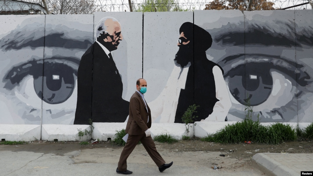 Seorang pria Afghanistan mengenakan masker melintas di depan foto utusan AS untuk Afghanistan, Zalmay Khalilzad, dan Mullah Abdul Ghani Baradar, pemimpin Taliban di Kabul, Afghanistan, 13 April 2020.