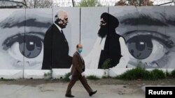 Seorang pria Afghanistan mengenakan masker melintas di depan foto utusan AS untuk Afghanistan, Zalmay Khalilzad, dan Mullah Abdul Ghani Baradar, pemimpin Taliban di Kabul, Afghanistan, 13 April 2020.