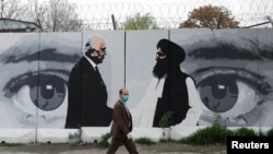 FILE - An Afghan man wearing a protective face mask walks past a wall painted with images of Zalmay Khalilzad, U.S. envoy in Afghanistan, and Mullah Abdul Ghani Baradar, the leader of the Taliban delegation, in Kabul, Afghanistan, April 13, 2020.