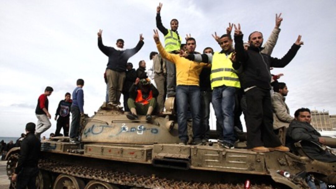 Para demonstran anti-pemerintah mengacungkan tanda kemenangan ("V" atau victory) dengan berdiri di atas sebuah tank militer di kota Benghazi (24/2).