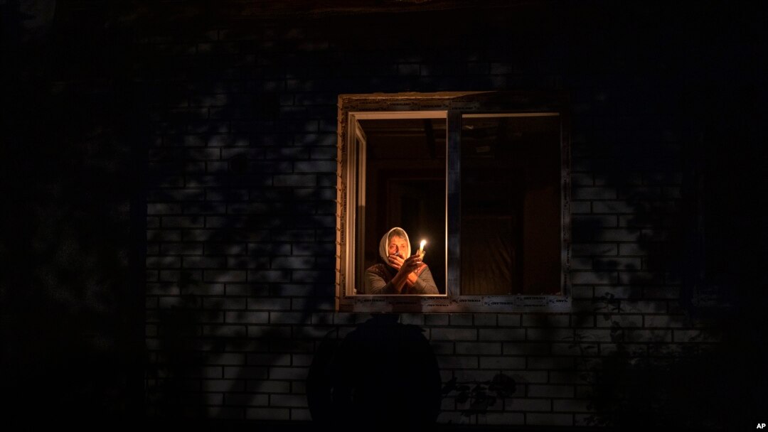 Catherine, 70, looks out the window while holding a candle for light inside her house during a power outage, in Borodyanka, Kyiv region, Ukraine, Thursday, Oct. 20, 2022. 
