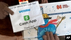 A vendor selling Street Sense holds up her sign saying she can accept donations from cashless apps like Venmo and CashApp, Wednesday, Dec. 6, 2023, in Washington. (AP Photo/Jacquelyn Martin)