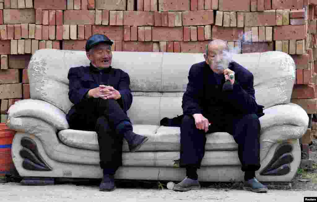 An elderly man (R) smokes a pipe as he chats with another man on a sofa in front of piles of bricks in Yongji, Shanxi province, China, April 11, 2015.