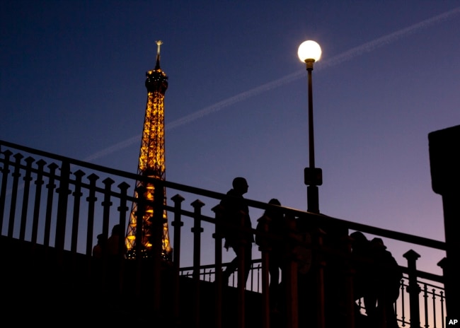 FILE - People walk on a bridge next to the Eiffel Tower in Paris, Wednesday Feb. 9, 2022. Lights on the Eiffel Tower will soon be turned off an hour earlier at night as part of an energy savings plan in the French capital, its mayor announced. (AP Photo/Rafael Yaghobzadeh, File)