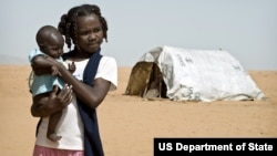 A young girl in Sudan holding a baby near a USAID tent in the Al Salam IDP (Internally Displace Persons) camp.