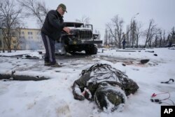 El cuerpo de un militar está cubierto de nieve mientras un hombre toma fotos junto a un vehículo militar de lanzacohetes ruso destruido, en las afueras de Kharkiv, Ucrania, el viernes 25 de febrero de 2022.