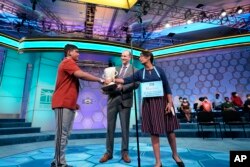 Harini Logan, 14, from San Antonio, Texas, right, and Vikram Raju, 12, from Aurora, Colo., left, wait on stage with Scripps CEO Adam Symson for results to be tabulated during the finals of the Scripps National Spelling Bee, Thursday, June 2, 2022, in Oxon Hill, Md. (AP Photo/Alex Brandon)