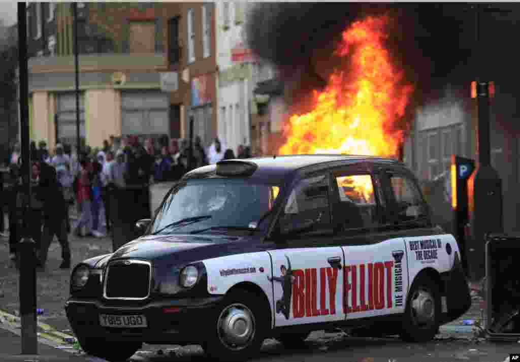 A car, behind the taxi, burns after it was set on fire by rioters in Hackney, east London, Monday Aug. 8, 2011. Youths set fire to shops and vehicles in a host of areas of London _ which will host next summer's Olympic Games _ and clashed with police in t