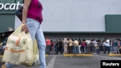 FILE - A woman carries bags with toilet paper rolls as people line up to buy staple items at a Makro supermarket in Caracas Aug. 4, 2015.