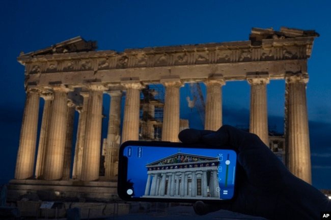 A woman holds up a mobile phone showing a digitally overlayed virtual reconstruction of the ancient Parthenon temple, at the Acropolis Hill in Athens, Greece on Tuesday, June 13, 2023. (AP Photo/Petros Giannakouris)