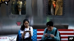 FILE - Shoppers sit on a bench with a decorated U.S. flag browsing their smartphones outside a fashion boutique selling U.S.-brand clothing at the capital's popular shopping mall in Beijing, Sept. 24, 2018.