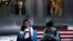 FILE - Shoppers sit on a bench with a decorated U.S. flag browsing their smartphones outside a fashion boutique selling U.S.-brand clothing at the capital's popular shopping mall in Beijing, Sept. 24, 2018.