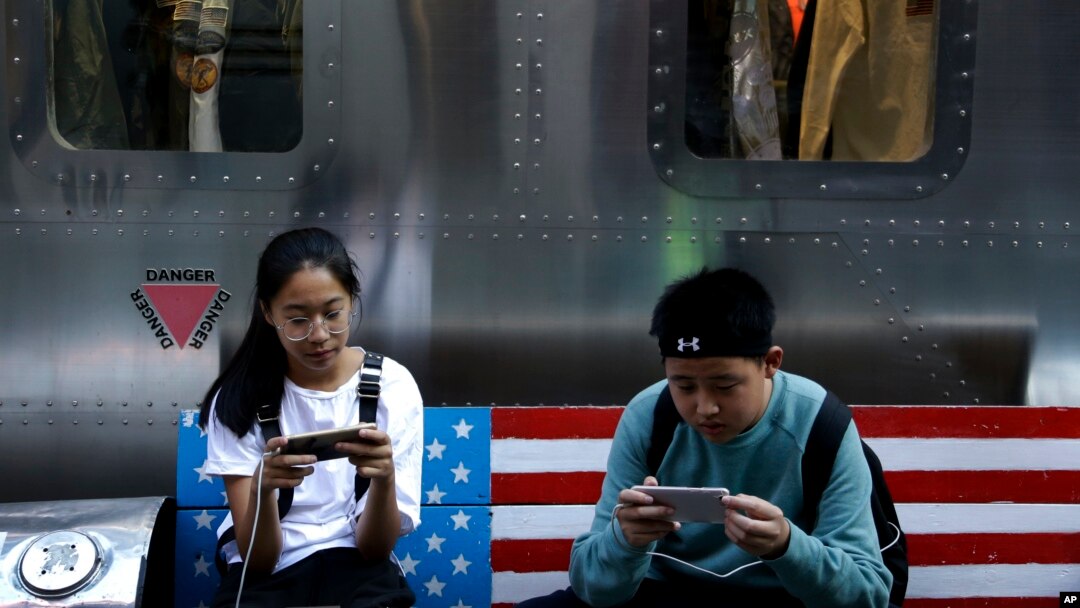 FILE - Shoppers sit on a bench with a decorated U.S. flag browsing their smartphones outside a fashion boutique selling U.S. brand clothing at the capital city's popular shopping mall in Beijing, Sept. 24, 2018.