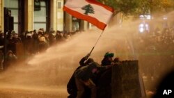 Anti-government protesters wave a Lebanese flag and hide behind a wood barrier from a water cannon as they clash with the riot police during a protest against the new government, near the parliament square, in Beirut, Lebanon, Jan. 22, 2020.