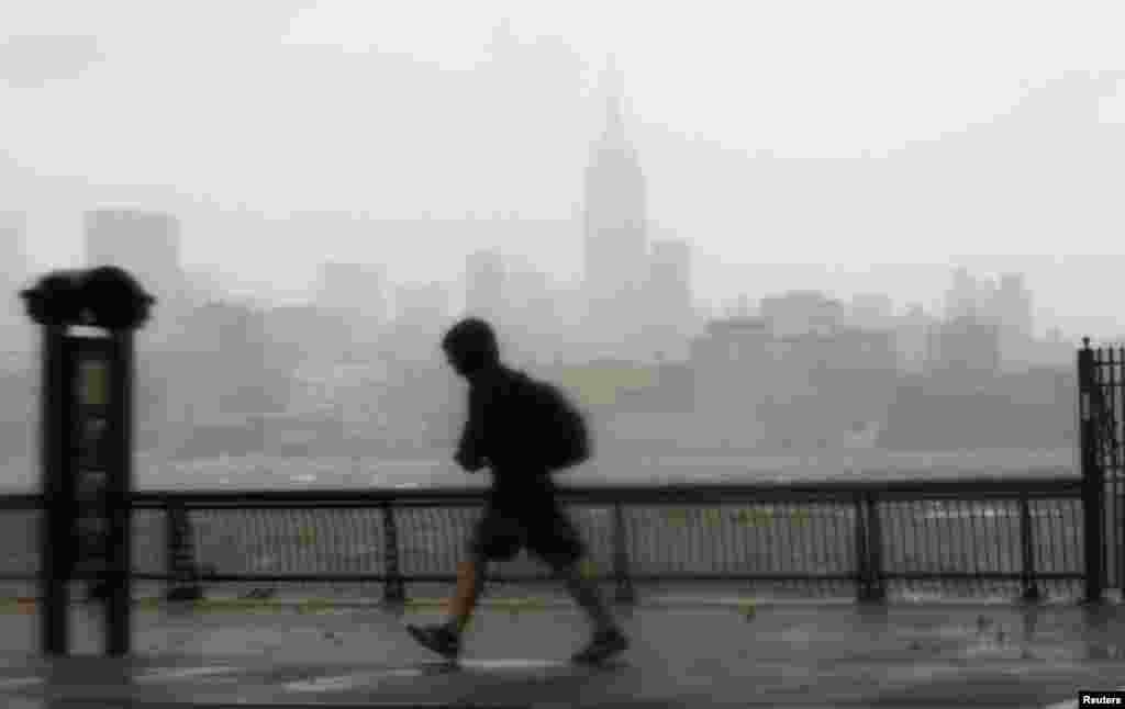 10月29日，一位男士冒着风雨走在新泽西州霍博肯的哈德逊河畔。对岸纽约市摩天大楼依稀可见。