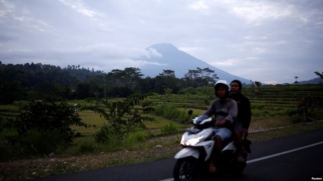 People ride a motorcycle past Mount Agung in the background, a volcano on the highest alert level, in Karangasem Regency, on the resort island of Bali, Indonesia, Sept. 24, 2017. 
